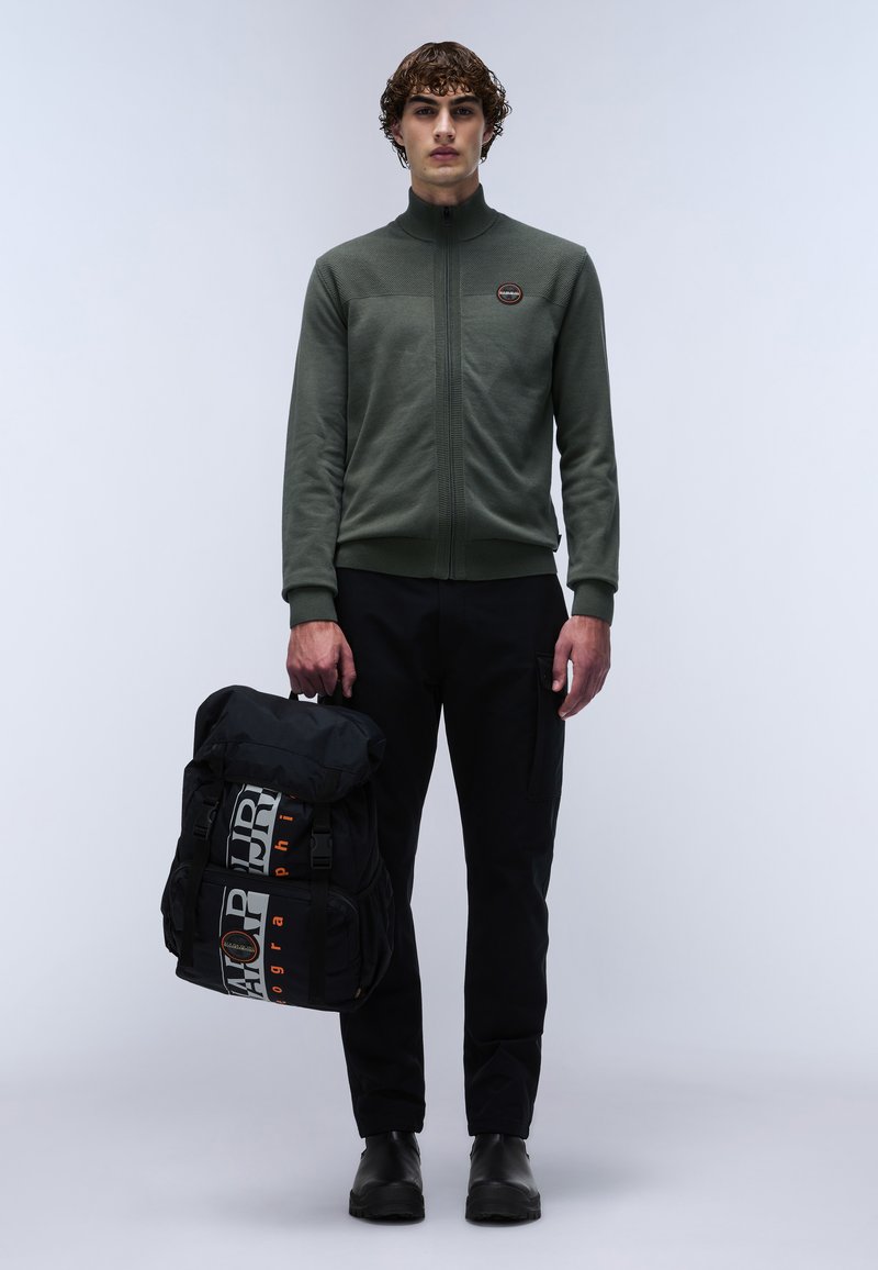 Veste zippée vert foncé avec un dessus texturé, pantalon cargo noir et un sac à dos noir avec des bretelles graphiques proéminentes et un logo.