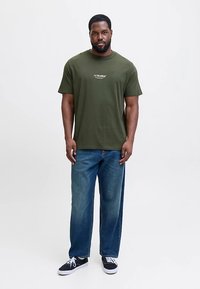 Maslinasto zelena pamučna majica kratkih rukava s bijelim logotipom. Uparena s ravnim trapericama plave boje i crnim tenisicama.