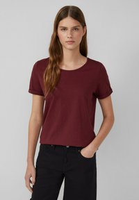 Bordeaux kortom shirt met een ronde halslijn, gemaakt van zachte stof. Heeft opgerolde mouwen en een ontspannen pasvorm, gecombineerd met zwarte broek.