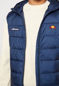 Tmavě modrá prošívaná vesta s osmistupňovým zipem, vybavená logovou nášivkou a prošívanou strukturou. Nošena přes krémovou mikinu s kulatým výstřihem.