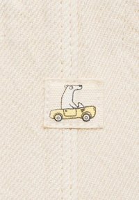 Un toppe in tessuto tessuto presenta un orso alla guida di una macchina cabriolet gialla. Lo sfondo è di un beige chiaro con un motivo diagonale testurizzato.