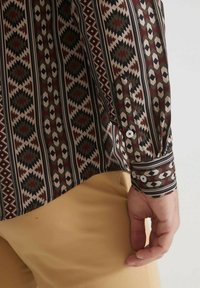 Chemise à manches longues avec un motif géométrique multicolore en marron, noir et beige, avec des poignets à boutons et une texture lisse.