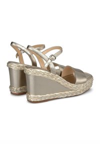 Sandales compensées avec des brides dorées métalliques et une plateforme en espadrille tissée. Comprend une sangle de cheville avec boucle et un design à bout ouvert.
