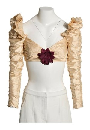 Crop-top beige à manches longues froncées, épaules bouffantes et ornement floral central rouge foncé, porté avec un pantalon blanc taille haute.