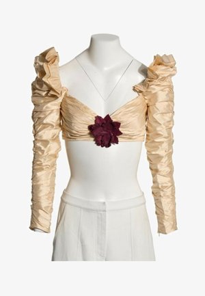 Crop-top beige à manches longues froncées, épaules bouffantes et ornement floral central rouge foncé, porté avec un pantalon blanc taille haute.
