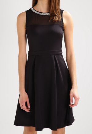 Czarna sukienka bez rękawów z przezroczystym panelem na górnej części klatki piersiowej oraz ozdobnymi srebrnymi dodatkami wokół dekoltu, dopasowaną talią i rozszerzaną spódnicą.