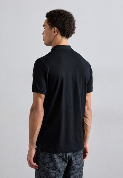 Svart polo t-shirt med korta ärmar, med en klassisk krage och slät yta, som bärs med mönstrade shorts, sedd från baksidan.
