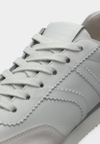 Λευκό δερμάτινο αθλητικό παπούτσι με υφασμάτινες λεπτομέρειες, ραφές σε ζιγκ ζαγκ και λεπτομέρειες από γκρι σουέντ. Διαθέτει επίπεδα κορδόνια και ενισχυμένη γλώσσα.