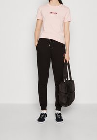 Różowa bawełniana koszulka z logo "ARMANI", czarne dresowe spodnie ze sznurkiem i czarna torba pikowana. Model nosi czarno-białe trampki.