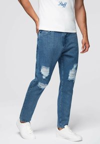 Jeans de mezclilla azul con un corte relajado, con desgastes y roturas en la parte delantera. Combinados con zapatillas blancas y una camiseta blanca básica.