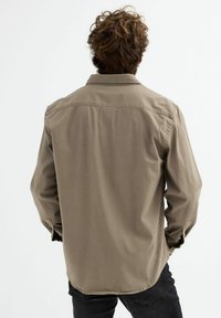 Camisa de manga larga color beige con un ajuste relajado, cuello abotonado y dobladillo curvo. La tela parece suave con detalles de costura mínimos.