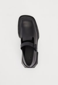 Black leather shoe with a square toe, adjustable strap, and a smooth texture. Features a slightly raised sole and minimalistic design.