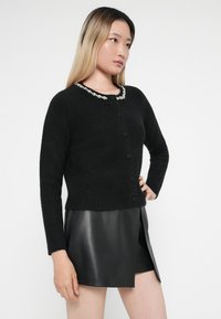 Cardigan nero in maglia testurizzata, con scollatura rotonda decorata da un bordo bianco e sei bottoni frontali.