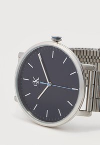 Ασημένιο ρολόι χειρός με μαύρο μινιμαλιστικό καντράν, ασημένιους δείκτες ώρας και λεπτών, μπλε δείκτη δευτερολέπτων και μεταλλικό λουρί πλέγματος.