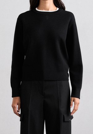 Femme portant un pull noir à manches longues avec des poignets et un ourlet côtelés, associé à un pantalon noir taille haute avec des poches à rabat latérales.