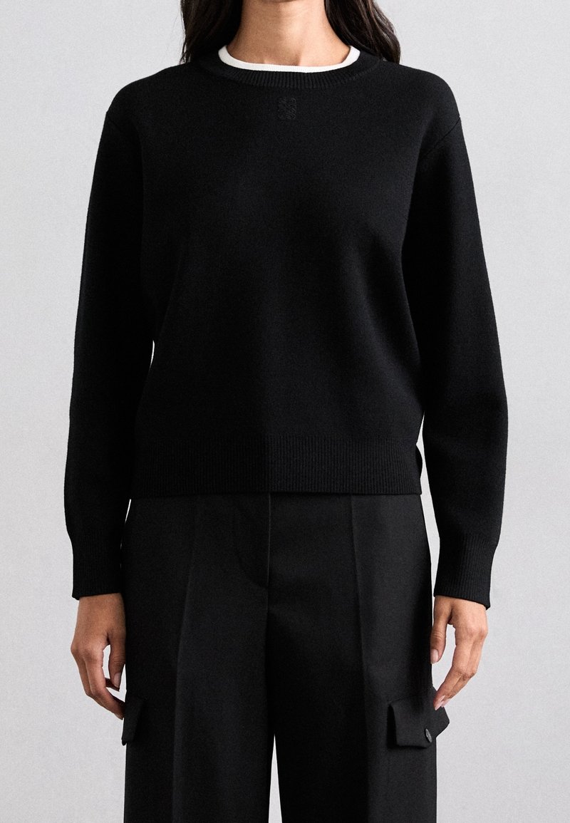Femme portant un pull noir à manches longues avec des poignets et un ourlet côtelés, associé à un pantalon noir taille haute avec des poches à rabat latérales.