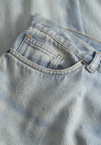 Detailný záber na svetlomodré denimové džínsy zobrazujúci predné vrecko, malé mincové vrecko, pútko na opasok a viditeľné stehové detaily.