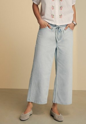 Frau trägt hellblaue Jeans mit weitem Bein und gebundenem Bund, weiße kurzärmelige Bluse mit Blumenstickerei und beigefarbene gewebte flache Schuhe.