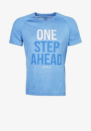 Hombre con camiseta azul "UN PASO ADELANTE", pantalones cortos deportivos negros y zapatos de correr grises, de pie sobre un fondo gris claro.
