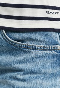 Top blanco de canalé con rayas horizontales en azul marino y el logo "GANT" encima de unos vaqueros de denim azul claro que presentan un bolsillo y costuras en contraste.