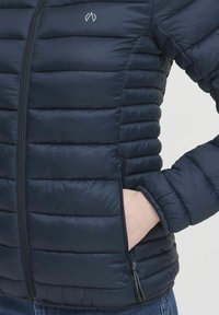 Námornícka páperová bunda so zipsom, horizontálnou prešívanou textúrou, bočnými vreckami a malým logom na hrudi. Mäkko a ľahký materiál.