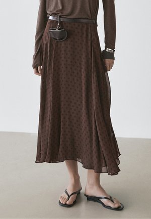 Persoon draagt een bruine midi-rok met polkadots, bijpassende top met lange mouwen, zwarte riemtas, zilveren polshorloge en zwarte teenslippers.