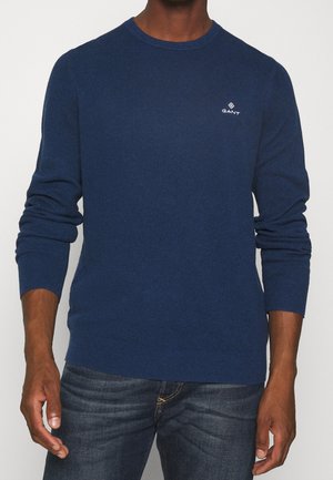 Pull en maille bleu marine avec un col rond, des poignets côtelés et un ourlet côtelé. Présente un petit logo blanc sur la poitrine gauche.