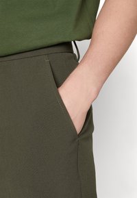 Personne portant un pantalon vert olive avec la main droite partiellement dans la poche avant, assorti à une chemise vert olive.