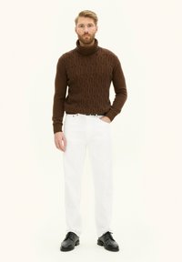 Maglione a collo alto lavorato a maglia marrone con motivo testurizzato, abbinato a jeans bianchi a gamba dritta e scarpe nere stringate. Sfondo neutro.