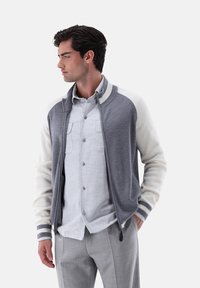 Graue Strickjacke mit weißen gerippten Ärmeln und Kragen, Reißverschluss und gestreiften Akzenten. Kombiniert mit einem hellgrauen button-up Hemd und hellen Hosen.