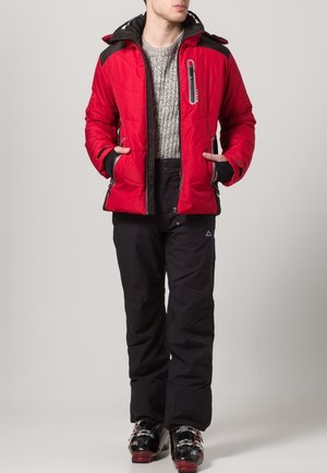 Veste isolée rouge avec une capuche noire et des accents, poches zippées, portée sur un pull en laine gris et un pantalon de ski noir avec des bottes.