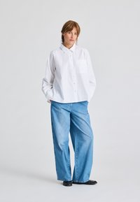 Camicia bianca a maniche lunghe con taschino, abbinata a jeans a gamba larga blu chiaro. Modello che indossa scarpe nere, in piedi davanti a uno sfondo grigio.