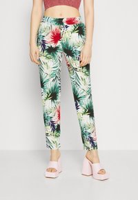 Femme portant un pantalon blanc avec un imprimé floral tropical coloré et des sandales à talons hauts roses, se tenant devant un fond uni.