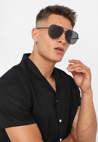 Ochelari de soare aviator negri cu lentile întunecate, cadru din metal, formă geometrică supradimensionată, purtați cu o cămașă neagră cu mânecă scurtă și butoni.