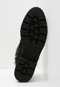 Semelle de botte noire à crampons profonds, en caoutchouc, avec des rainures et motifs pour l'adhérence. Vue latérale montrant des accents en fourrure et une construction robuste.
