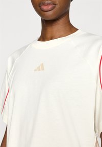 Kreemjas t-särk raglaanvarrukatega, millel on helepruun logo ja punased aktsendid õmbluste ääres. Sile tekstuur, vaba lõige.