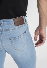 Jeans de mezclilla azul claro con un diseño ajustado. Incluyen un parche de cuero marrón etiquetado como "GIANNI KAVANAGH" y dos bolsillos traseros con costuras visibles.