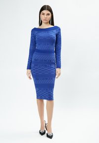 Abito blu a costine con motivo testuale "INFLUENCER", maniche lunghe, silhouette aderente, abbinato a scarpe con tacco nere con dettagli decorativi.