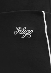 Κοντινή λήψη μαύρου υφάσματος με λευκή κεντημένη επιγραφή "Hugo" και κάθετη λευκή ρίγα στη δεξιά πλευρά.