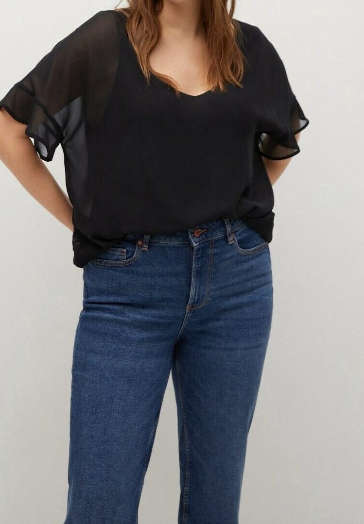 Femme portant un chemisier noir transparent à manches courtes rentré dans un jean bleu taille haute, debout devant un fond clair uni.