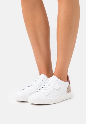 Bílé nízké tenisky se šněrováním a růžovými a béžovými akcenty, nošené na bosých nohách na jednobarevném světlém pozadí.