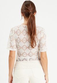 Haut en dentelle blanche avec un motif de diamants complexe, manches courtes, ourlet festonné et une texture semi-transparente, associé à un pantalon de couleur claire.