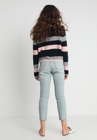 Žena s dlouhými kudrnatými vlasy stojí čelem ke zdi, obléká pruhovaný svetr, světle modré zkrácené džíny a bílé slip-on boty.