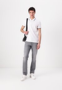 Biely polo tričko s čiernymi detailami na golieri, šedé džínsy a biele tenisky. Model drží malú čiernu crossbody tašku. Jednoduché pozadie.