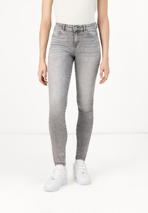 Persoon die lichtgrijze, hooggetailleerde skinny jeans en witte sneakers draagt, staand tegen een effen witte achtergrond.