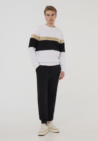 Felpa bianca con strisce orizzontali nere e beige e testo, abbinata a pantaloni jogger neri e sneakers beige. Tessuto di cotone liscio.