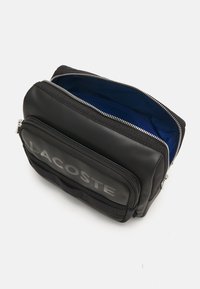 Černá taška se zipem s hladkým povrchem, vybavená přední kapsou a logem "LACOSTE", podšitá modrou látkou. Viditelné detaily ze stříbrných komponentů.