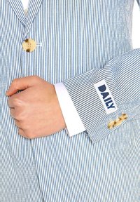 Zilā un baltā svītraina žakete ar vienu pogu, baltu krekla piedurkni un tagu ar uzrakstu "DAILY" zilā krāsā uz piedurknes.