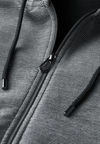 Felpa grigia con zip, caratterizzata da un tessuto testurizzato, cerniera nera e cordini, con un primo piano della cerniera e dei dettagli del materiale.