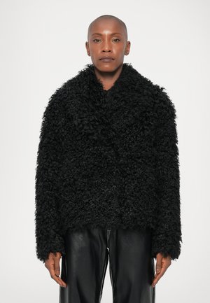 Fekete műszőrme dzseki, texturált, bolyhos felülettel, laza szabással és széles gallérral. Fekete bőrnadrággal párosítva.